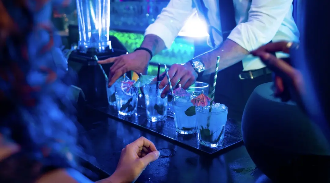 Bartender serving cocktails behind the bar in a busy nightclub environment.